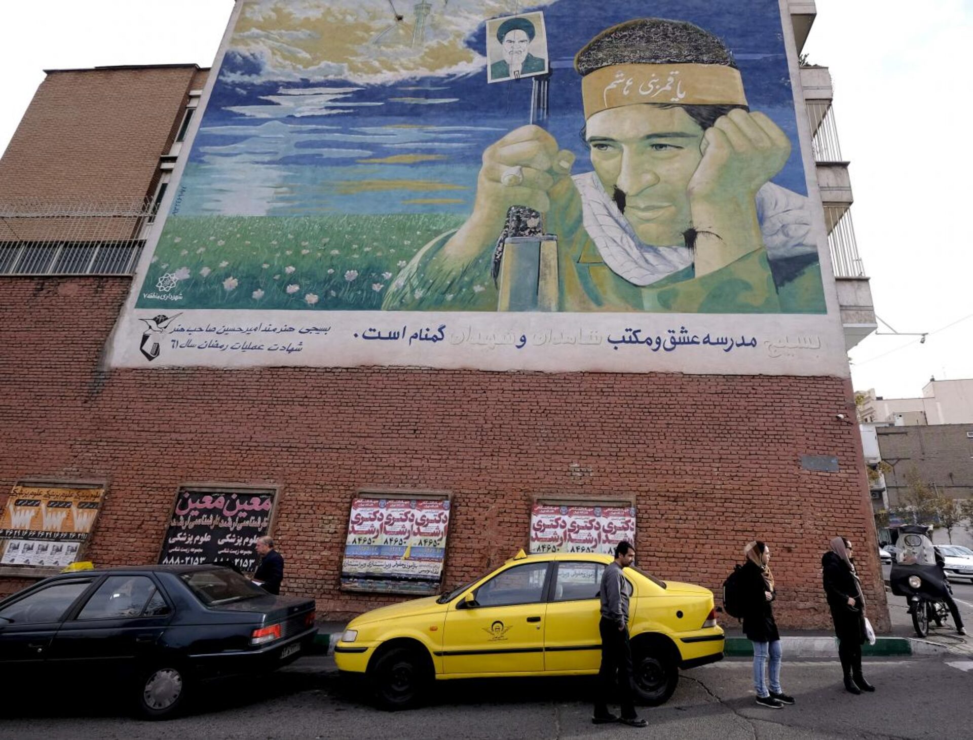 People standing under a worn-out mural depicting a soldier killed in the Iraq-Iran war, Tehran, Iran, Dec. 2015