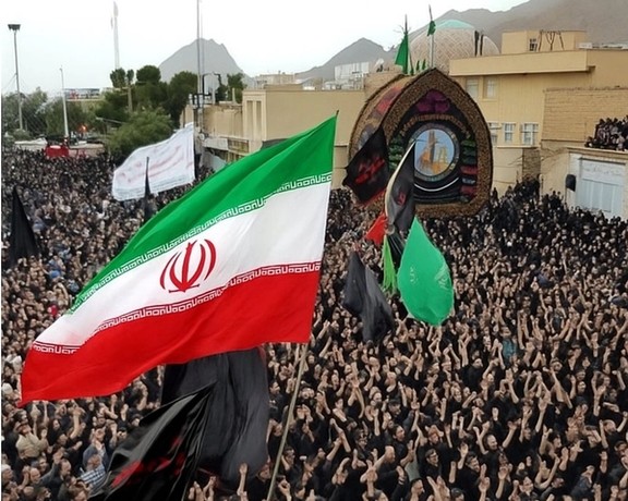A large group of mourners gather in a religious ceremony waving the Iranian flag, July 2025