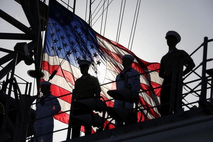 A US flag flutters in the background as members of the Marines and the Navy stand on a deck of the USS Bataan, a Navy Wasp-class amphibious assault ship, deployed to the Persian Gulf in July.
