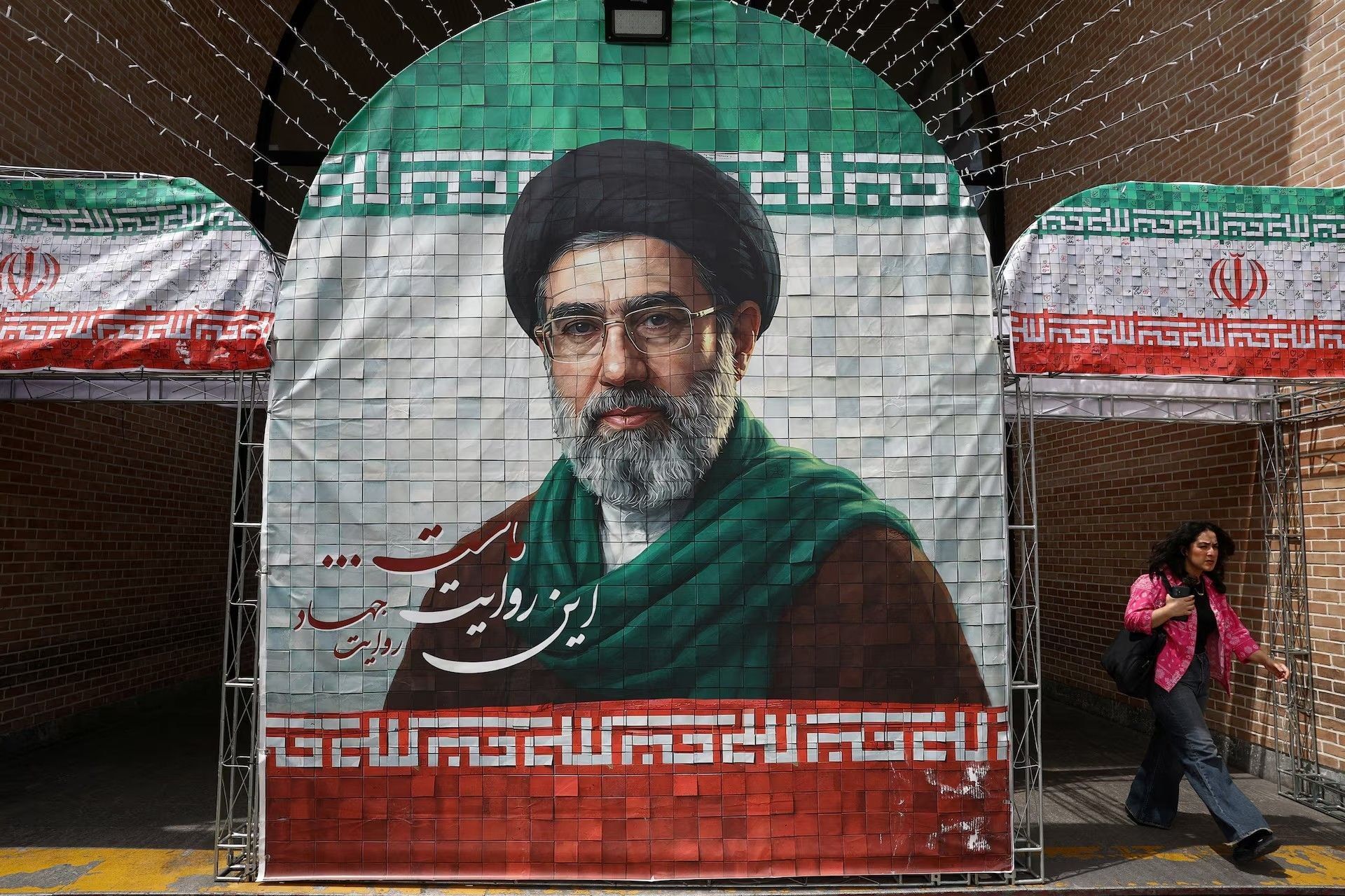A large portrait of Mojtaba Khamenei is displayed alongside Iranian flags at a public venue.