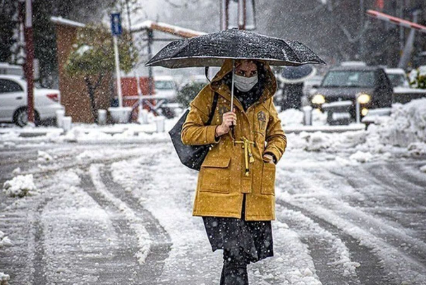 A masked pedestrian walks under heavy snowfall, shielding themselves with an umbrella as winter intensifies, causing widespread closures and disruptions across Iran.