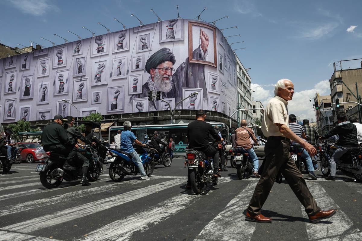 A man crosses a street in Tehran as motorcyclists pass beneath a giant billboard depicting Iran’s slain supreme leader Ali Khamenei, May 6, 2026