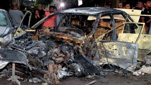 People stand near a damaged vehicle at the site of a bomb blast, outside the Sayeda Zeinab shrine city south of the Syrian capital Damascus, Syria July 27, 2023.