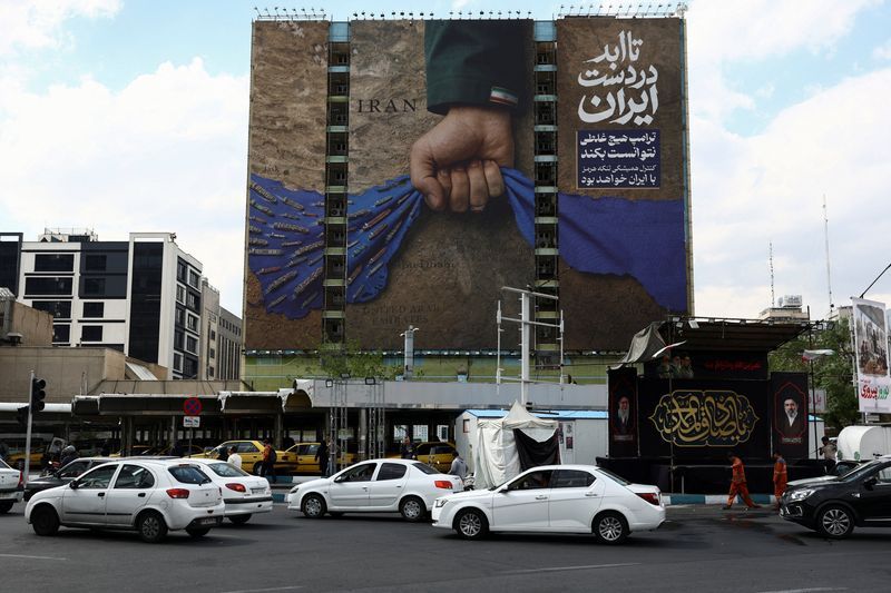 A huge billboard in Tehran shows a clenched hand gripping a blue cloth shaped like the Strait of Hormuz, symbolizing Iran’s hold over the strategic waterway, with Persian text reading “Forever in Iran’s hands,” displayed above a busy intersection in the capital.