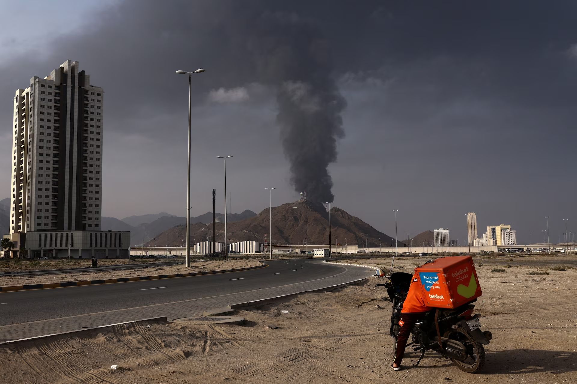A person stands next to a motorcycle as smoke rises in the Fujairah oil industry zone following a fire caused by debris after interception of a drone by air defenses, according to the Fujairah media office, United Arab Emirates, March 3, 2026.     