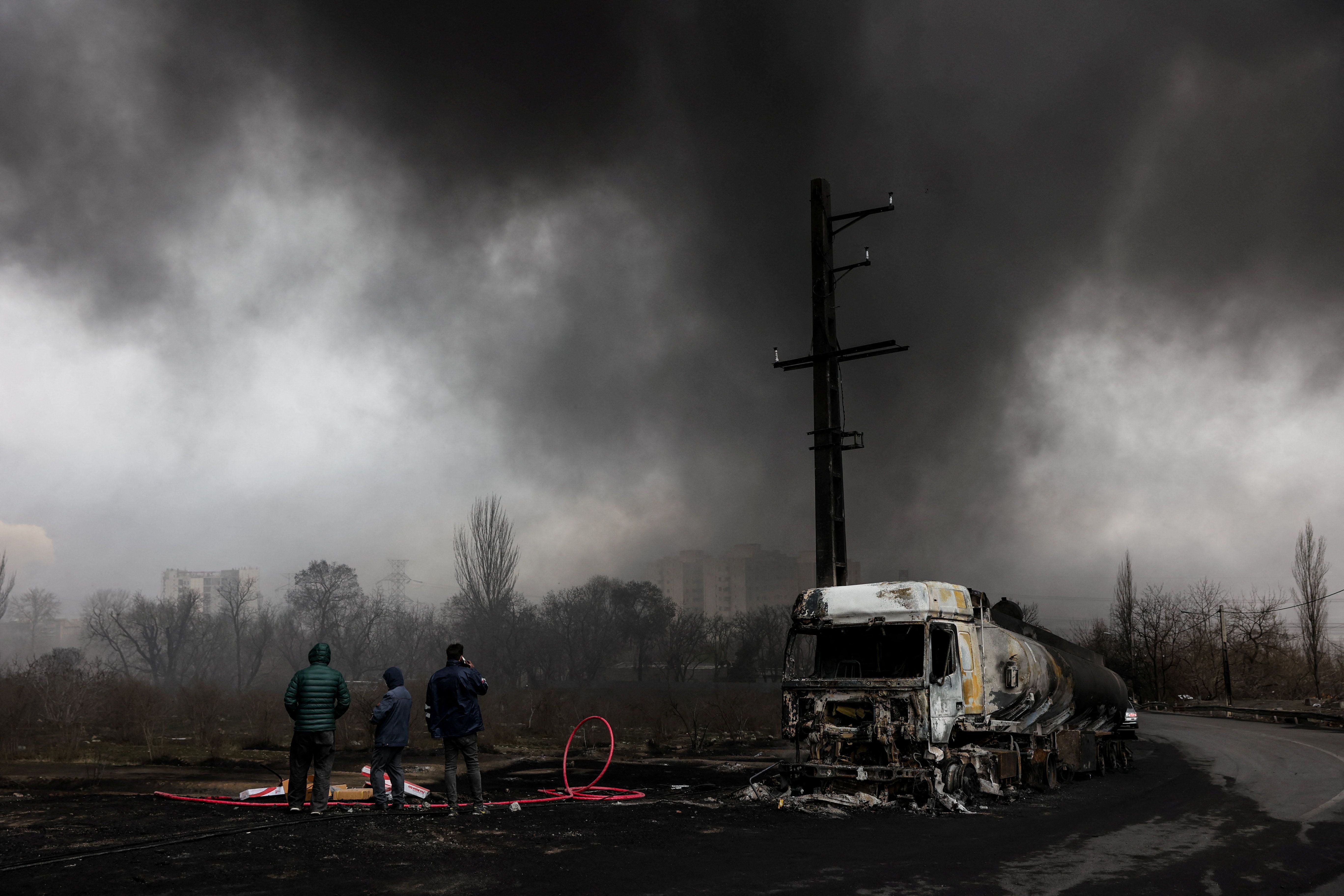 People stand near a destroyed vehicle as smoke rises after a reported strike on Shahran fuel tanks, amid the US-Israeli conflict with Iran, as seen through a window, in Tehran, Iran, March 8, 2026. 